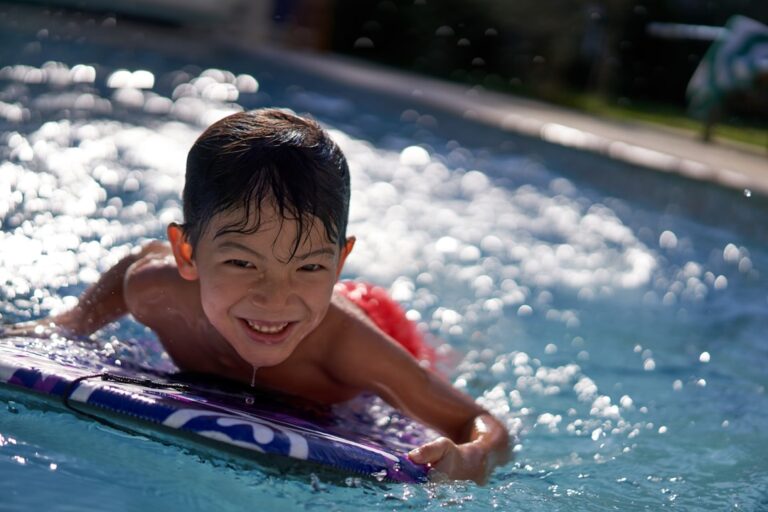 Children playing and splashing in a backyard pool with wide entry steps and a shaded seating area nearby.