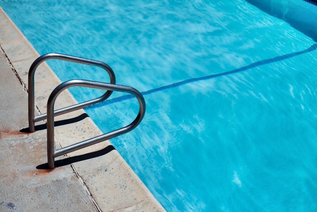 A person skimming leaves from a backyard pool surrounded by lush green landscaping and clear water.
